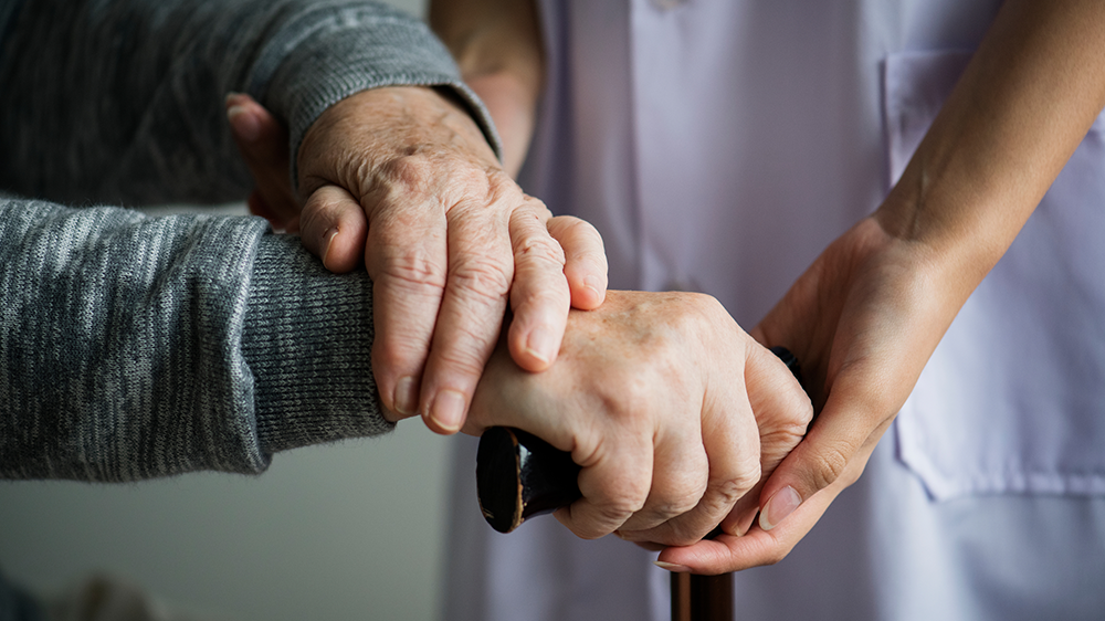 Symbolbild für den Bereich Altenhilfe: zwei Hände einer älteren Person stützen sich auf einen Gehstock, zwei Hände einer jüngeren Person in Pflegekleidung stützen die ältere Person