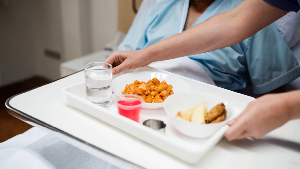 Eine Pflegekraft bringt einem Patienten ein Tablett mit Essen ans Bett.